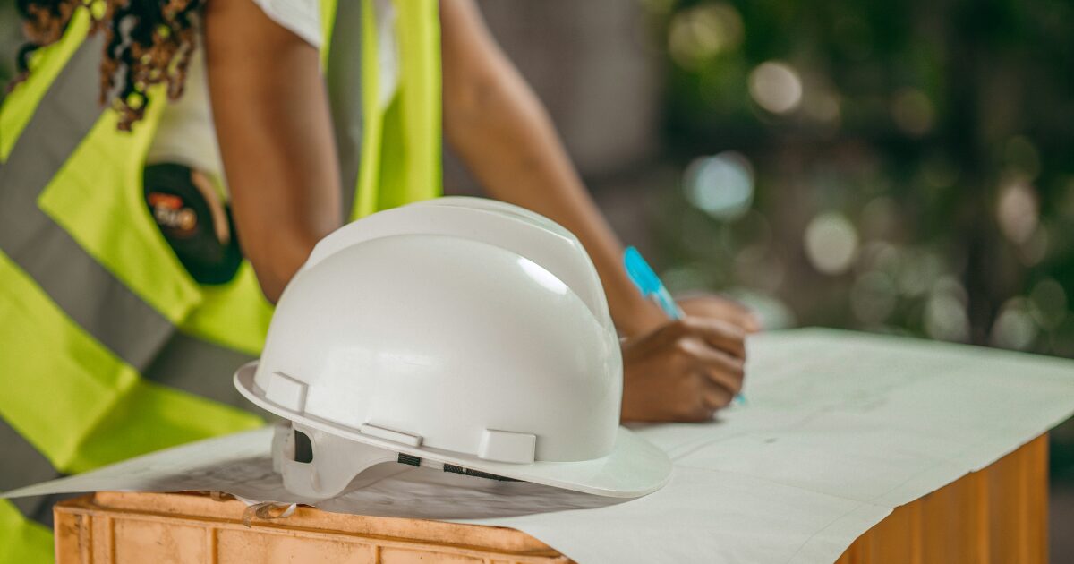 235 An African female construction worker holding a pen and writing on a paper. The desk has a hard hat and paper.