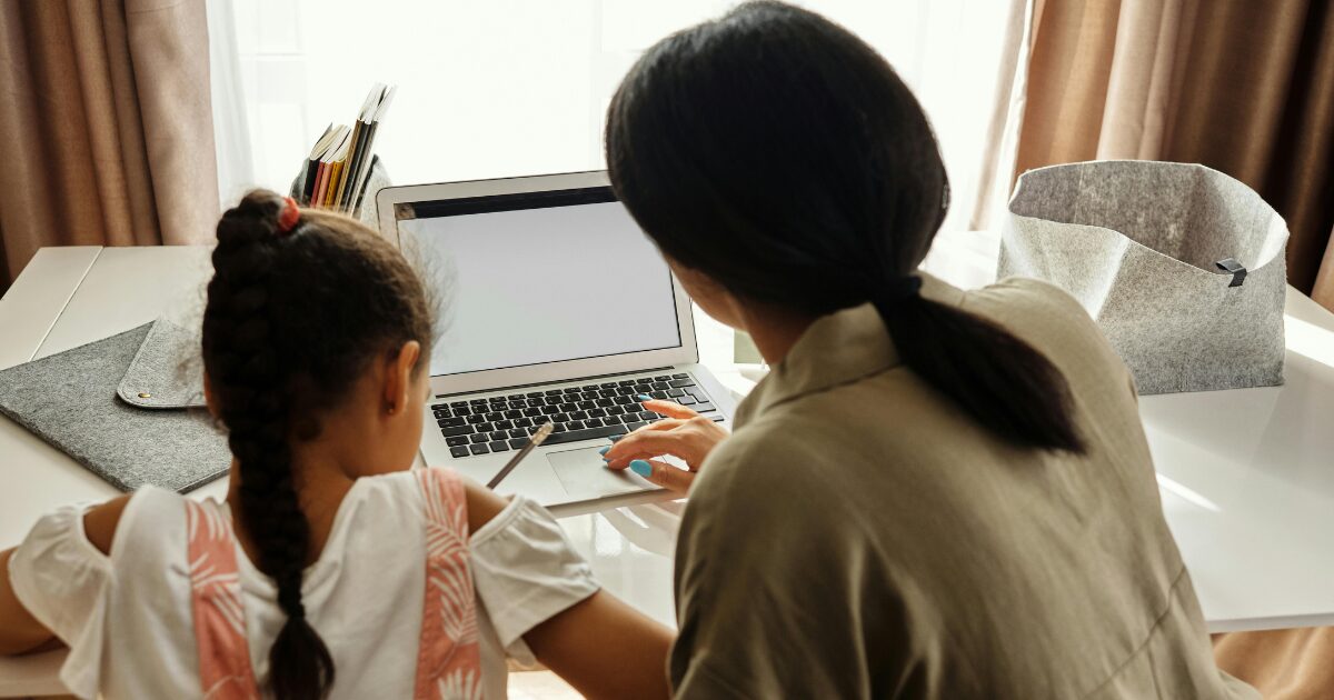 237 A Southeast Asian mother and her Southeast Asian daughter sitting in front of a laptop. The Southeast Asian mother with black hair is using the laptop while her Southeast Asian daughter with black hair is writing something down. They are facing away from the camera.