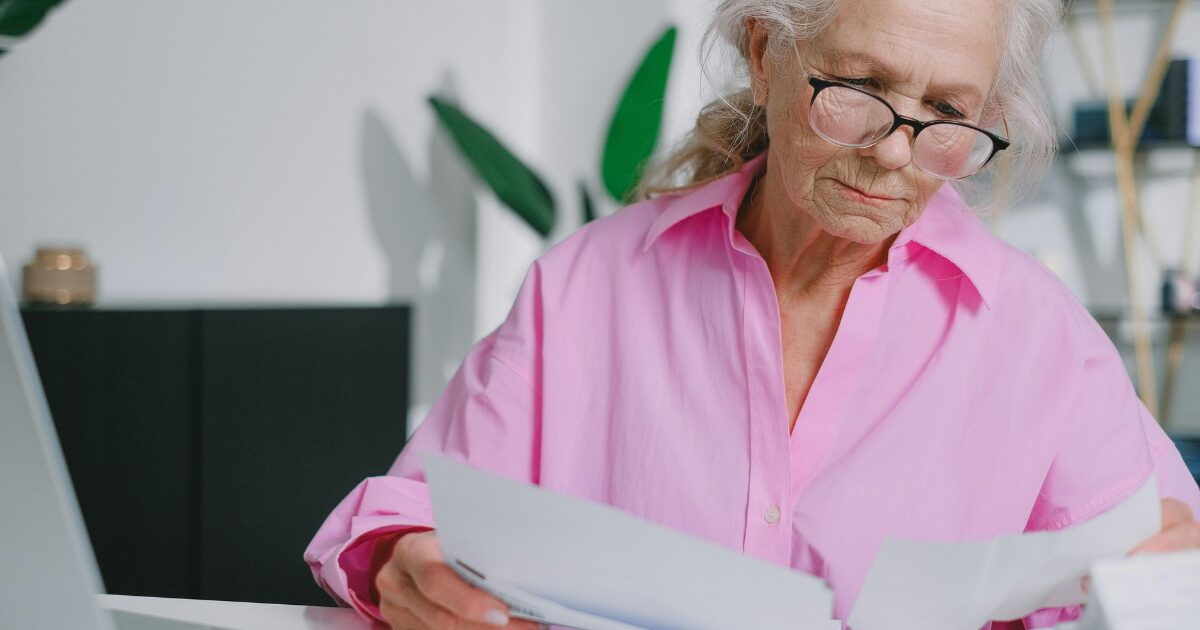 An elderly Caucasian woman wearing a pink blouse, reading over some documents