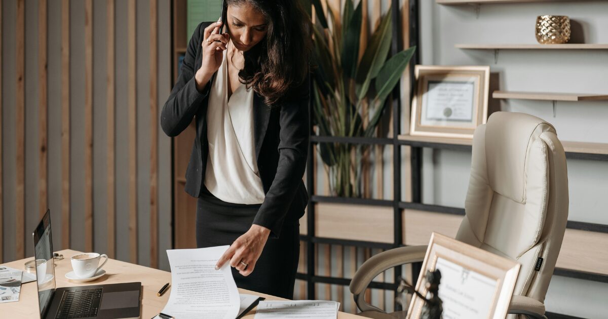 A Black woman in a black blazer, standing over a desk, while looking at a document as she’s on a phone call