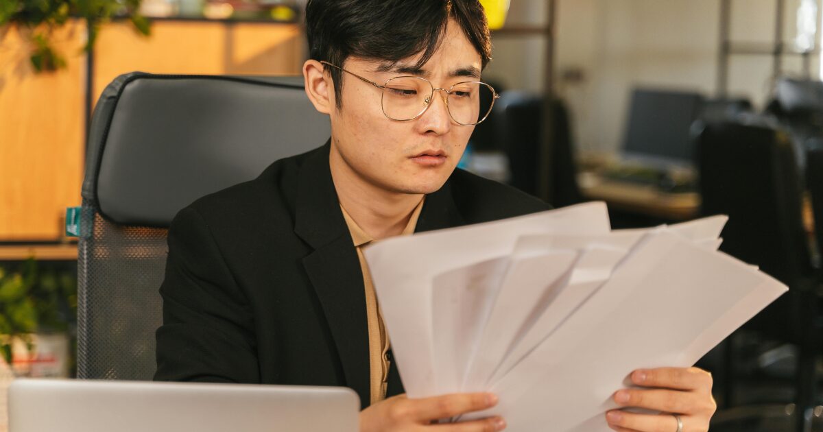 An East Asian man wearing a black blazer, sitting at a desk, reading several documents in his hands