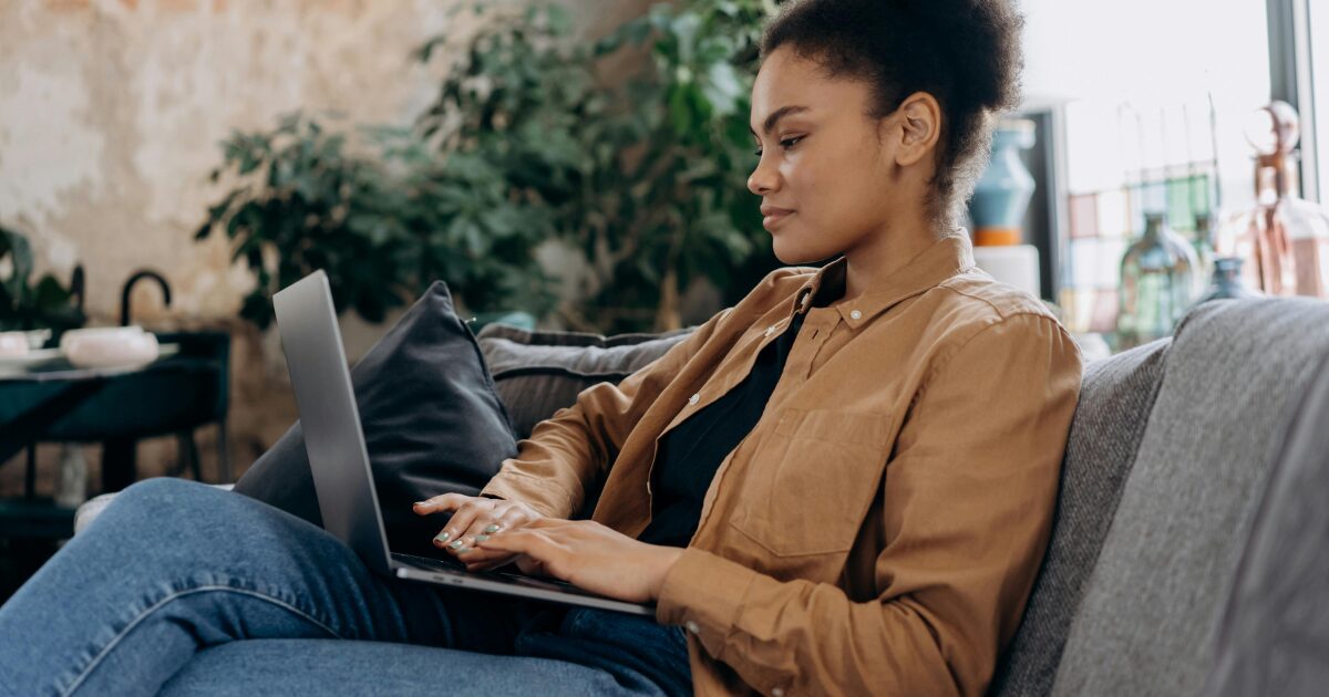 255 A Black woman in a brown jacket, sitting on a grey sofa while typing on a laptop on her lap