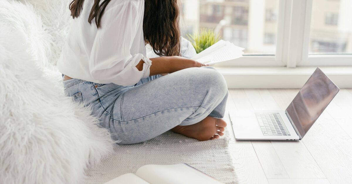 256 A faceless woman in a white blouse, sitting cross-legged on the floor, while holding documents in her hand. A laptop is on the floor in front of her.
