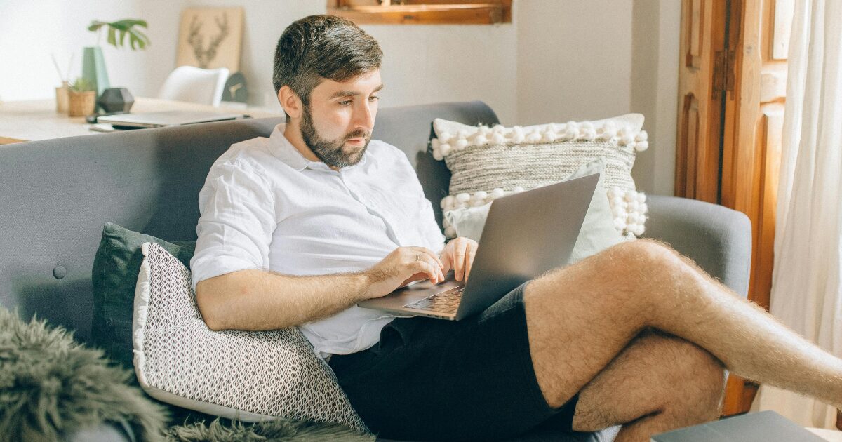 258 A Caucasian man in a white polo, sitting on a grey sofa while typing on a laptop on his lap.