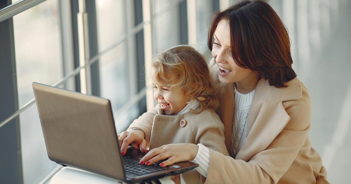 260 A Caucasian woman and a young Caucasian girl, both smiling while looking at a laptop. The mom is typing and both are wearing tan-coloured coats.