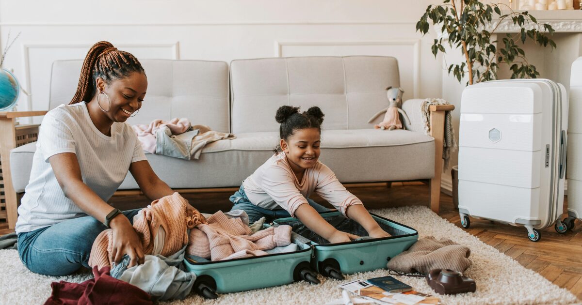 262 A Black woman and a young Black girl smiling while packing clothes into a suitcase. The woman is wearing a white shirt while the girl is wearing a beige top.