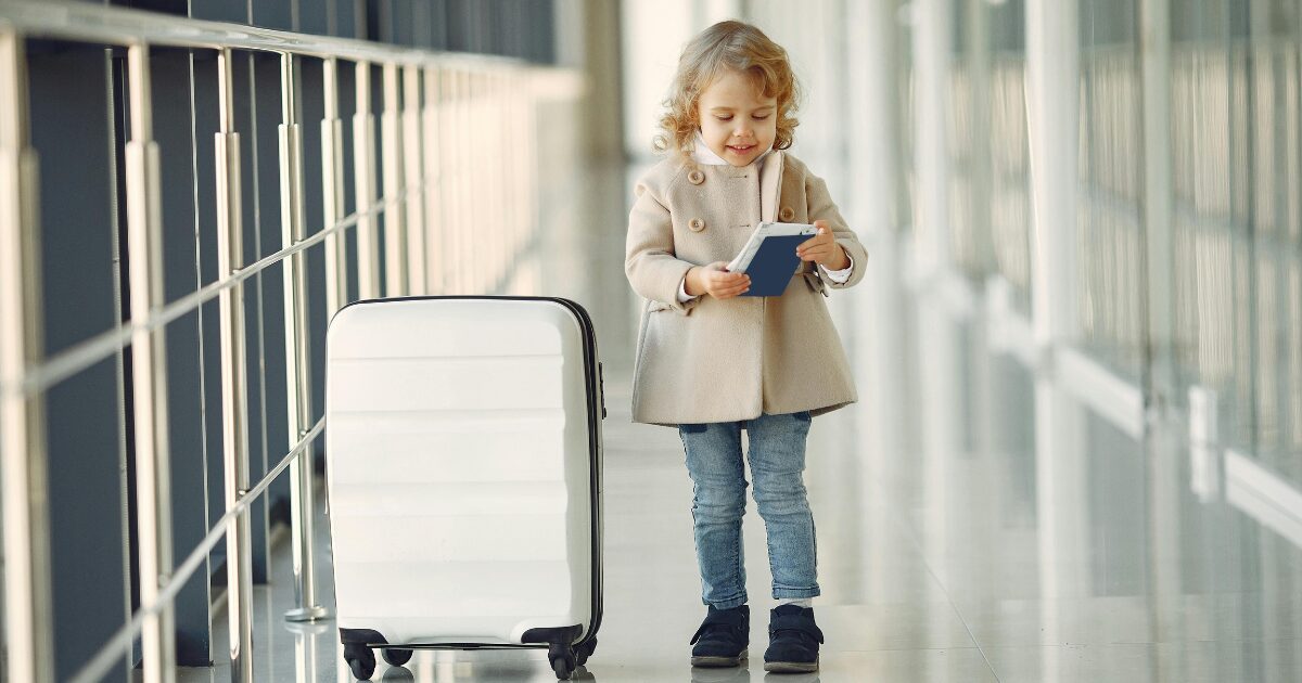 264 A young Caucasian girl smiling while looking down at a passport in her hands. She is standing beside a white suitcase.