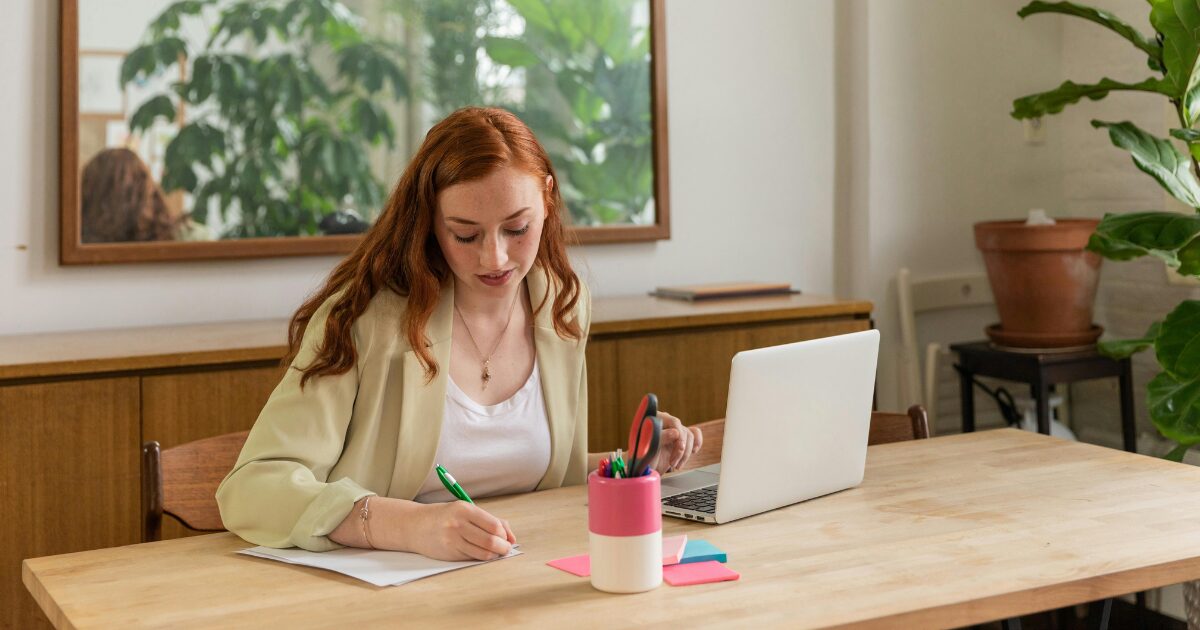 270 A Caucasian woman in a cream-colored blazer writing on a piece of paper while sitting at a desk in front of a laptop