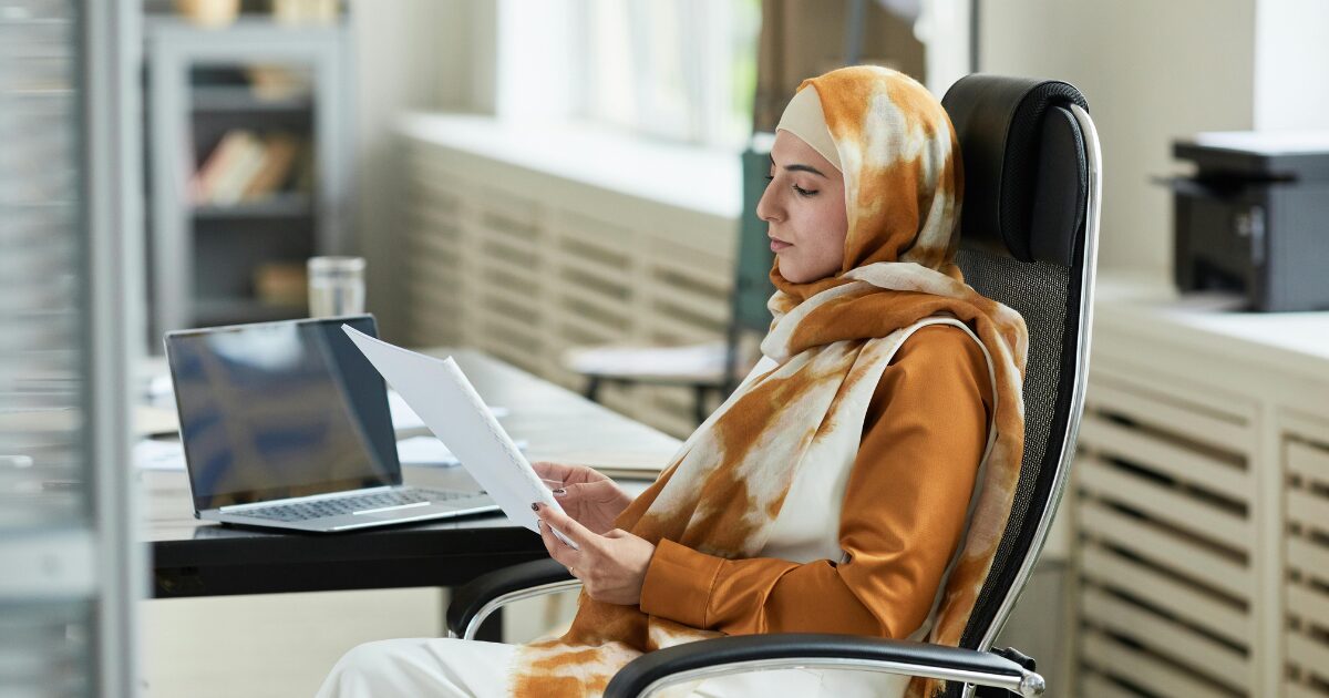 273 A Middle Eastern woman wearing a hijab reading a document while sitting in an office chair