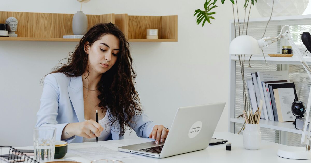 274 A Caucasian woman in a blue blazer, sitting at a white desk in front of a laptop, while pointing at a document on the table with a pen.