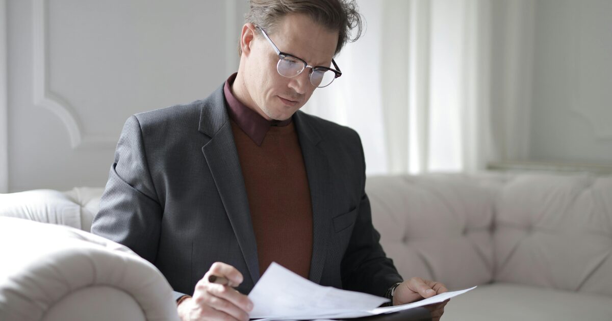 276 A Caucasian man wearing a black blazer, reading a document while sitting on a bright gray sofa.