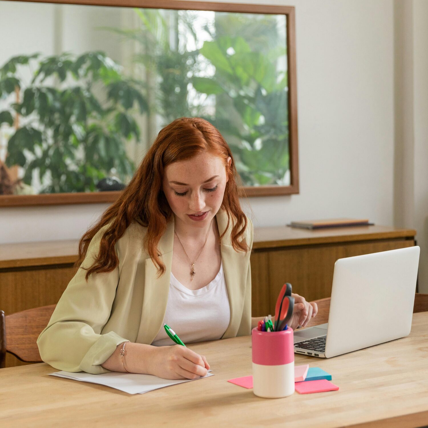 A Caucasian woman in a cream-colored blazer writing on a piece of paper while sitting at a desk in front of a laptop