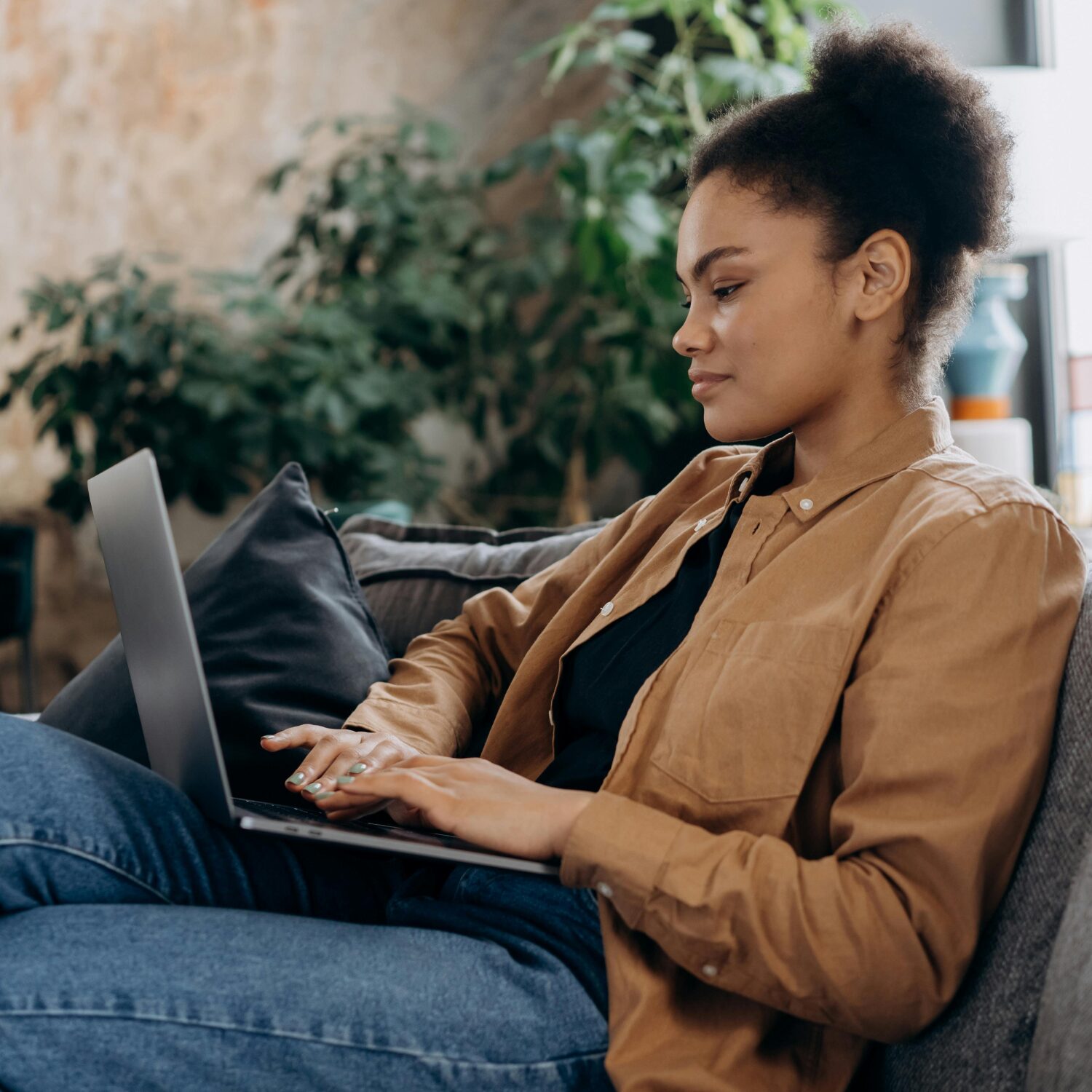 A Black woman in a brown jacket, sitting on a grey sofa while typing on a laptop on her lap