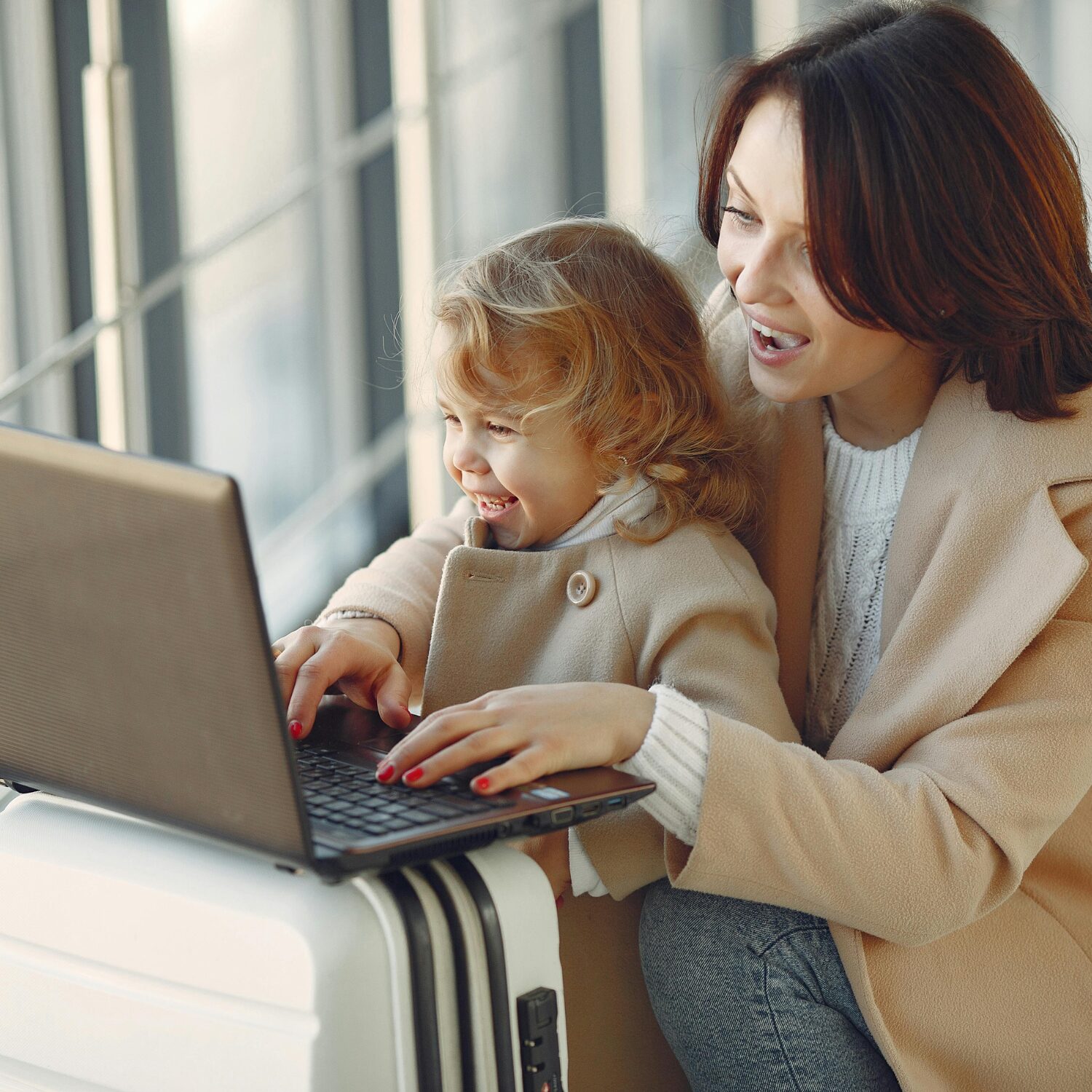A Caucasian woman and a young Caucasian girl, both smiling while looking at a laptop. The mom is typing and both are wearing tan-coloured coats.