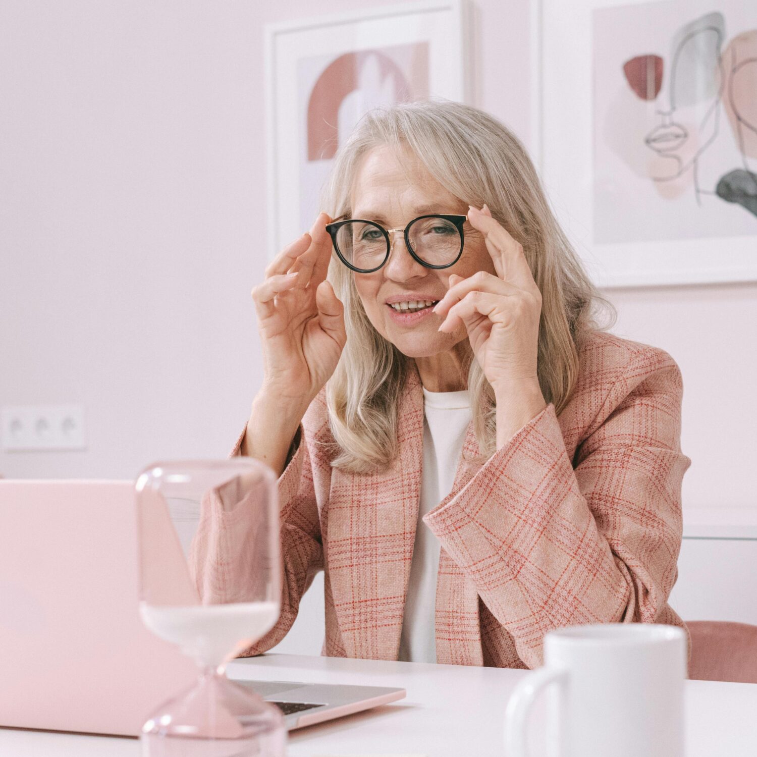 An elderly Caucasian woman sitting at a desk, looking at a laptop with a curious smile. She's wearing eye-glasses and a pink plaid blazer