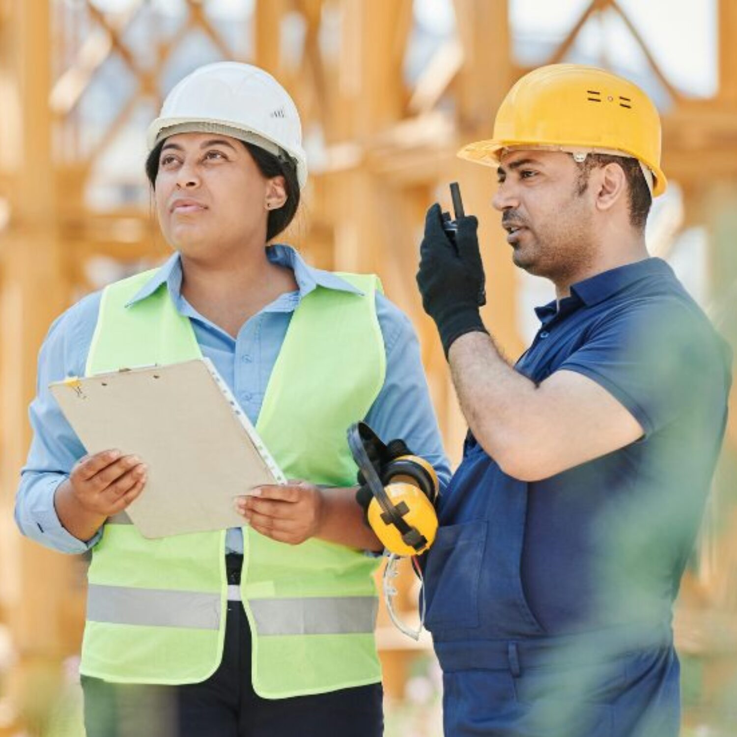A Black woman and Latino man standing on a construction site. The woman is holding a clipboard and both are wearing hard hats.