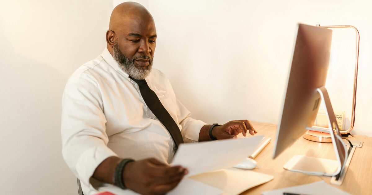 Black man wearing a white button-white short, sitting a desk, looking at a document in his hand