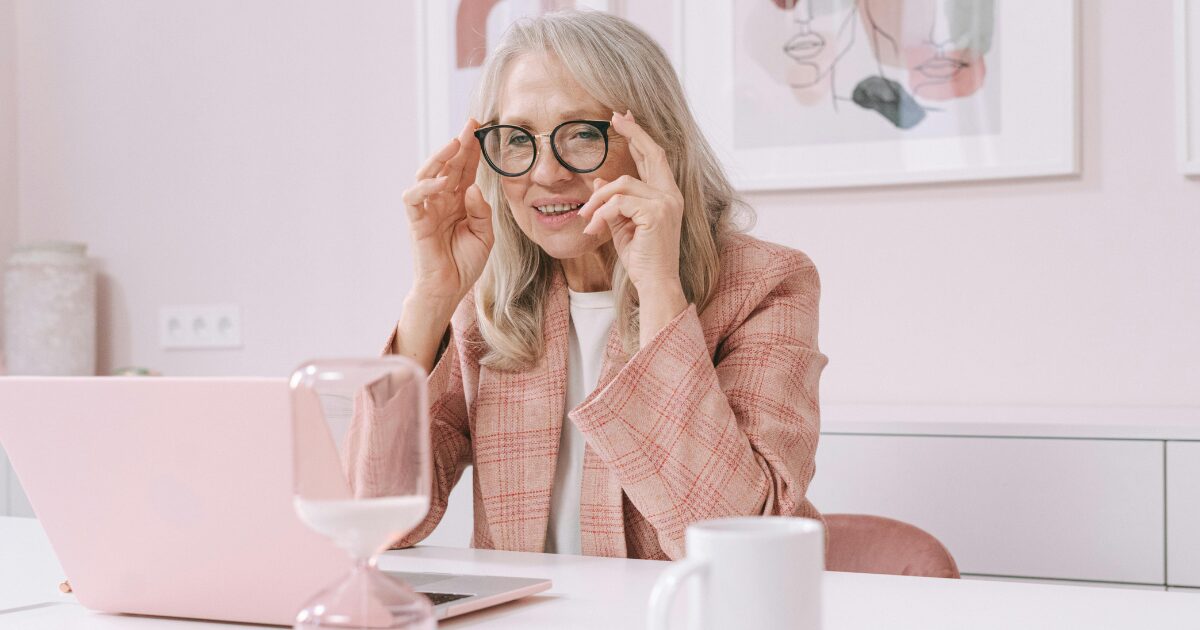 An elderly Caucasian woman sitting at a desk, looking at a laptop with a curious smile. She's wearing eye-glasses and a pink plaid blazer