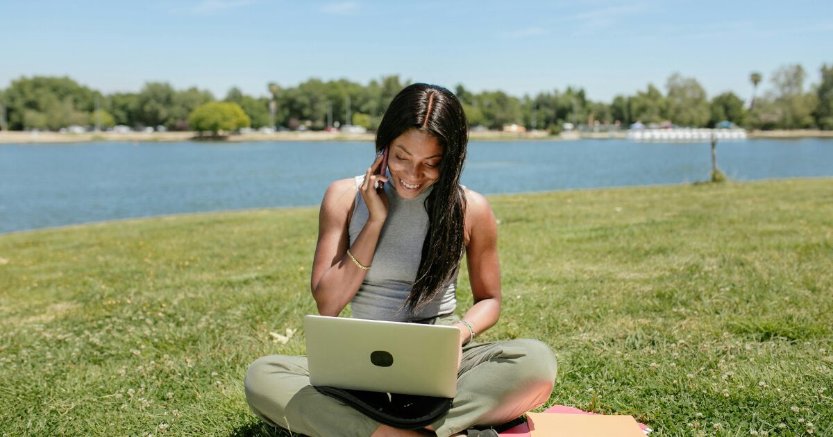 287 A Black woman sitting cross-legged on the grass with a laptop on her lap. The woman is smiling while she’s on a phone call.