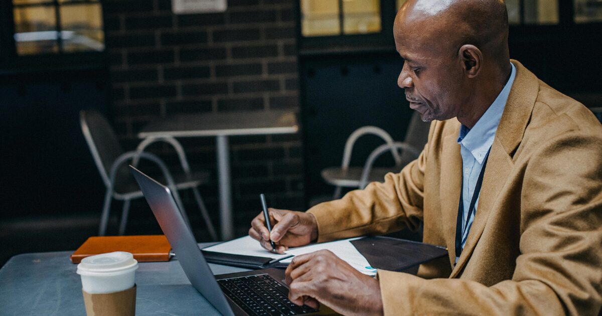 289 A Black man wearing a brown blazer writing on a document while sitting in front of a laptop