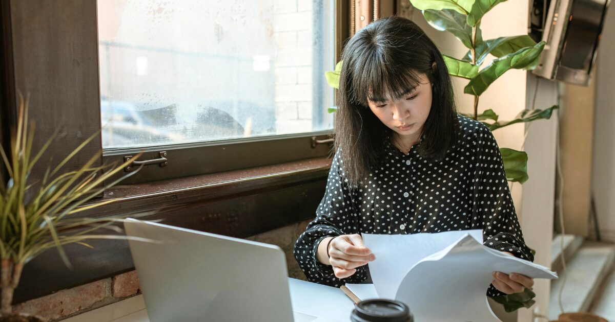 292 An East Asian woman wearing a black polka-dot button-up blouse flipping through pages of a document while sitting in front of a laptop.