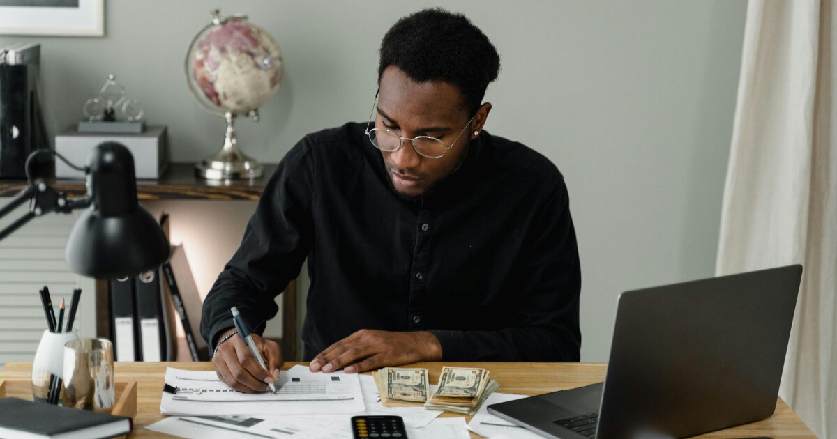 293 A Black man wearing a black button-up shirt while writing on a document. The man is sitting in front of a laptop.