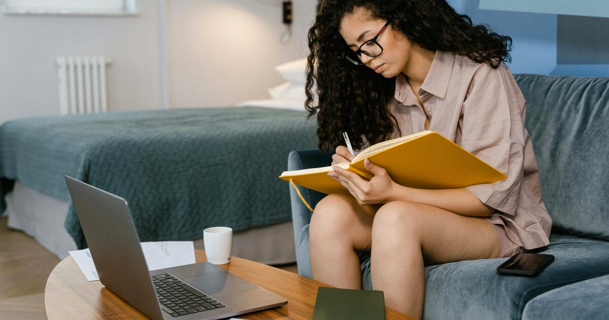 A Black woman wearing a tan blouse sits on a grey sofa, writing in a notebook with a laptop open in front of her