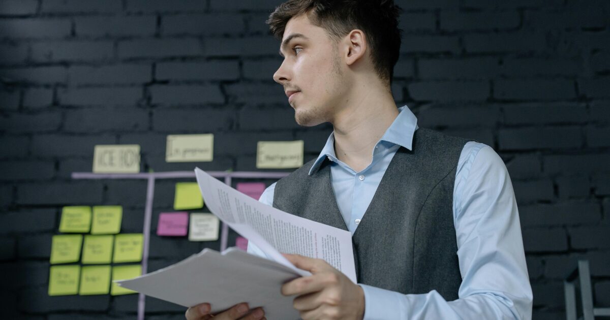296 A Caucasian man wearing a sky-blue long-sleeve shirt and gray vest faces sideways while holding documents