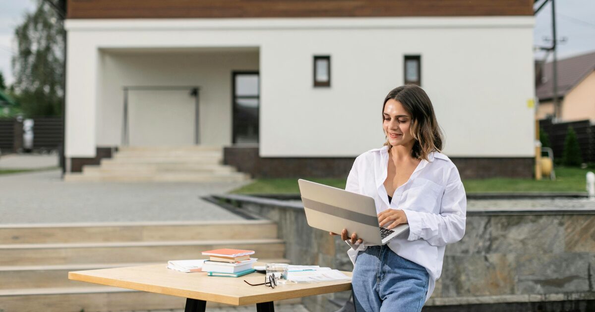 A Caucasian woman wearing a white blouse, smiling as she holds an open laptop while standing outdoors.