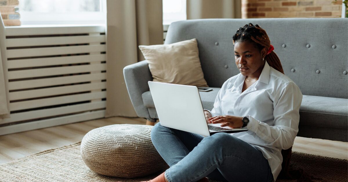 A Black woman wearing a white blouse, sitting on the floor with a laptop on her lap.