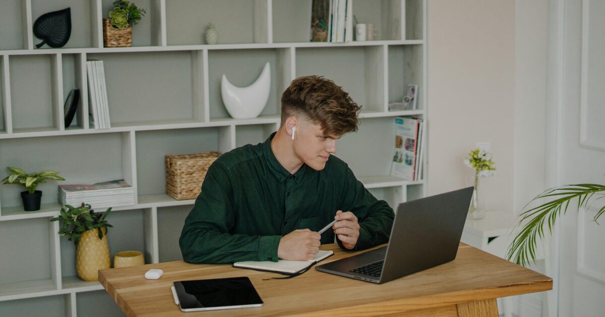 A Caucasian man wearing a dark green long-sleeved shirt, holding a pen while sitting in front of an open laptop.