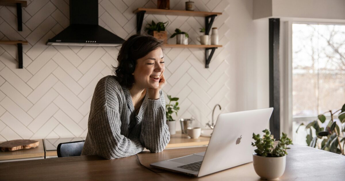 A Caucasian woman wearing a gray sweater, smiling while sitting in front of a laptop.