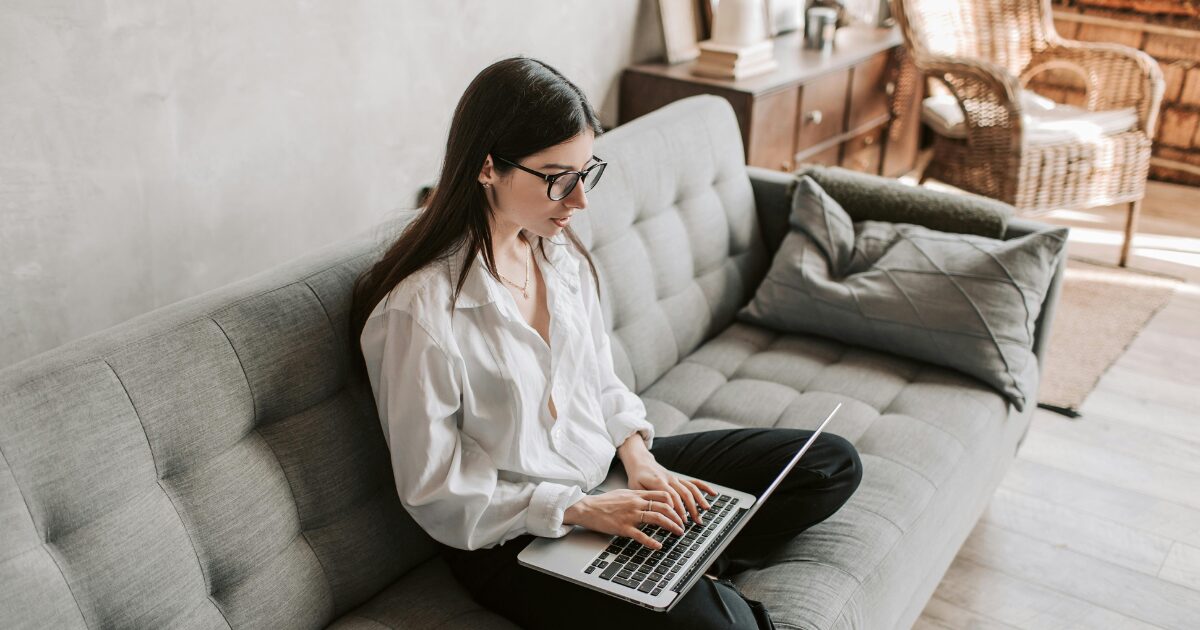 A Caucasian woman wearing a white long-sleeved blouse, sitting cross-legged on a gray sofa with a laptop on her lap.
