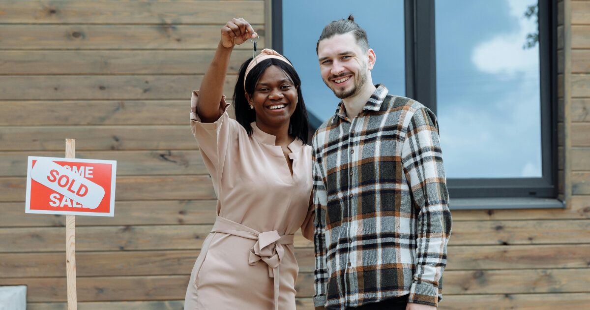 308 A young Black woman wearing a soft pink dress, posing with a Caucasian man in a plaid shirt. She is holding a key and both are smiling, standing in front of a house.