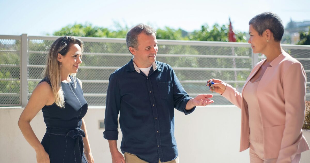 309 A Caucasian woman in pink blazer handing keys to a Caucasian man wearing a black buttoned shirt and another Caucasian woman wearing a black dress.