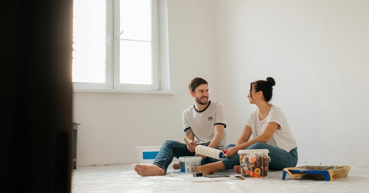 312 A Caucasian man and woman both wearing white shirts, sitting on the floor while surrounded by painting materials.