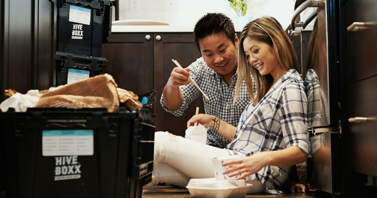 313 An East Asian man and woman both wearing checkered shirts, sitting on the floor and smiling while eating food.