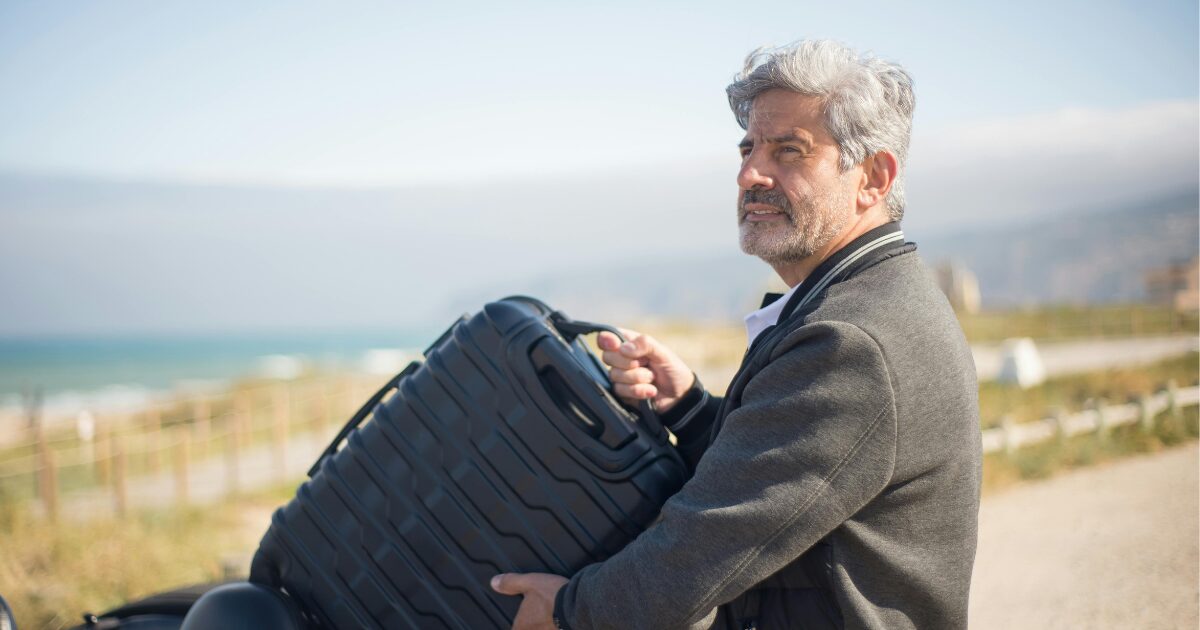 318 An Elderly Caucasian man wearing a gray jacket carrying a black travel luggage outdoors.
