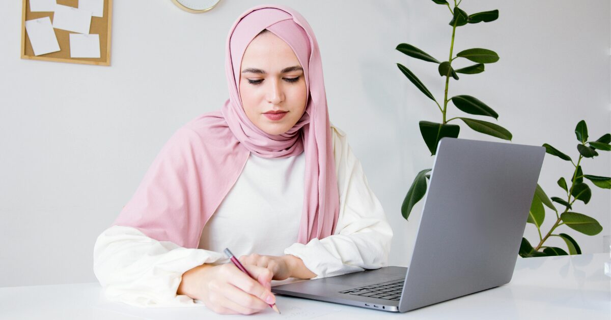 319 A Caucasian woman wearing a white long sleeve blouse and a pink hijab writing something down while sitting in front of a laptop.