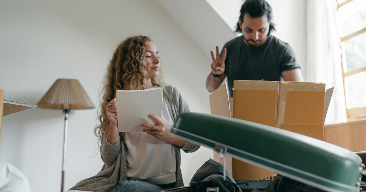 322 A Caucasian woman wearing a gray cardigan holds documents while a Caucasian man in a black T-shirt rummages through a box behind her.