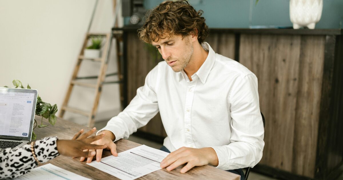 A Caucasian man wearing a white long sleeved shirt looking at a document on the table