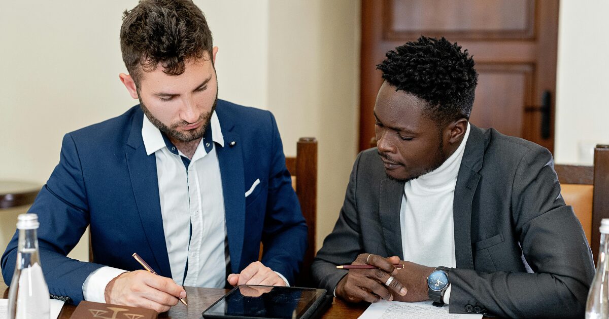 A Caucasian man wearing a navy blue business suit and a Black man wearing a black business suit sitting together while looking at a tablet
