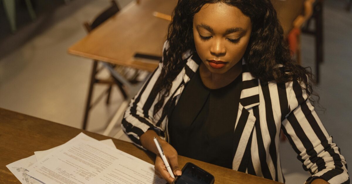 A Black woman wearing a black and white striped blazer looking at her phone while holding a pen on her right hand