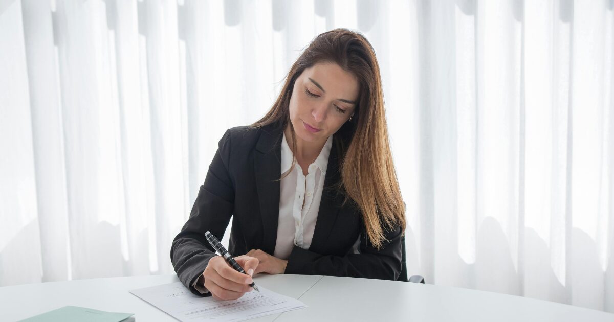 A Caucasian woman wearing a black blazer writing on a piece of paper.