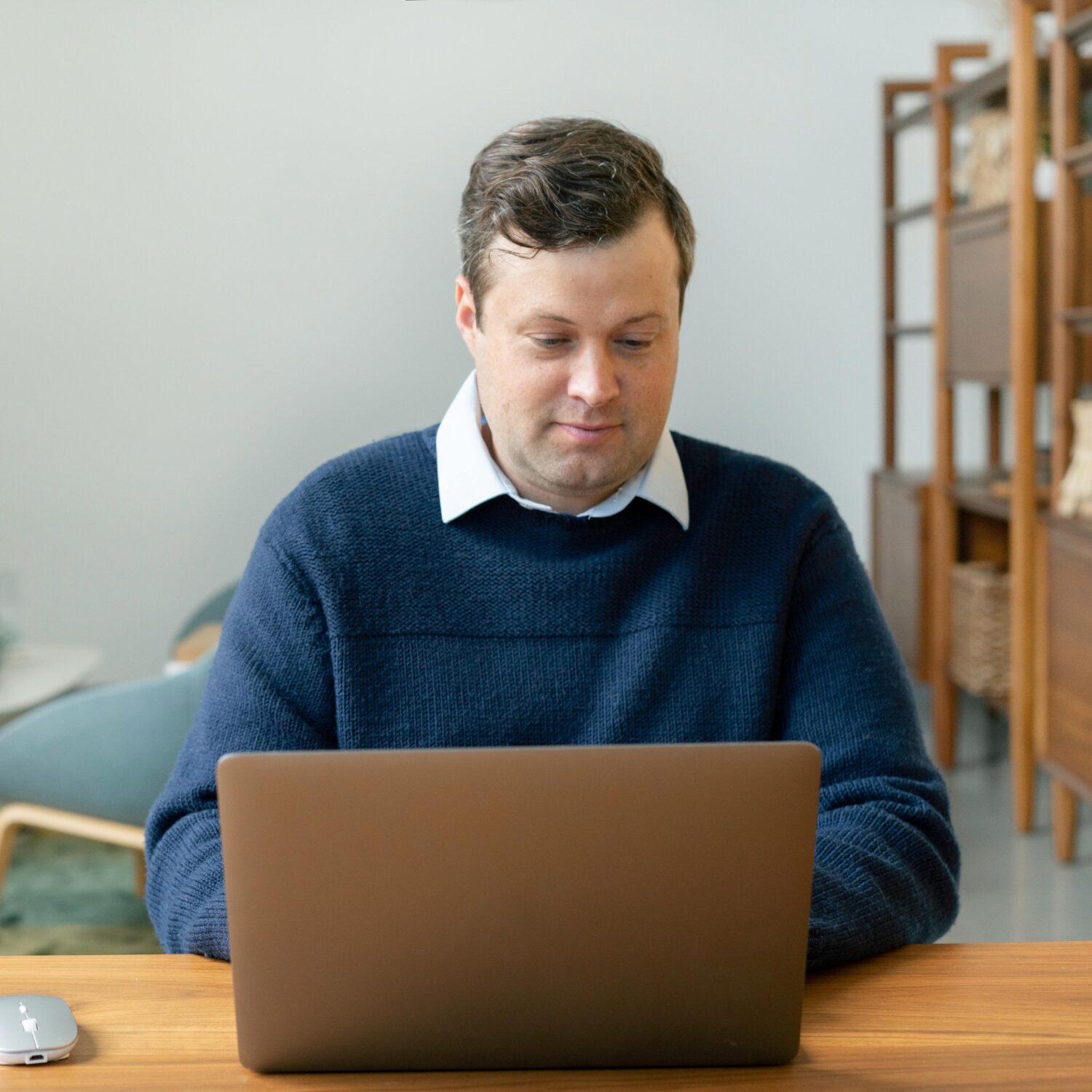 A Caucasian man wearing a blue collared sweater smiling while sitting in front of his laptop