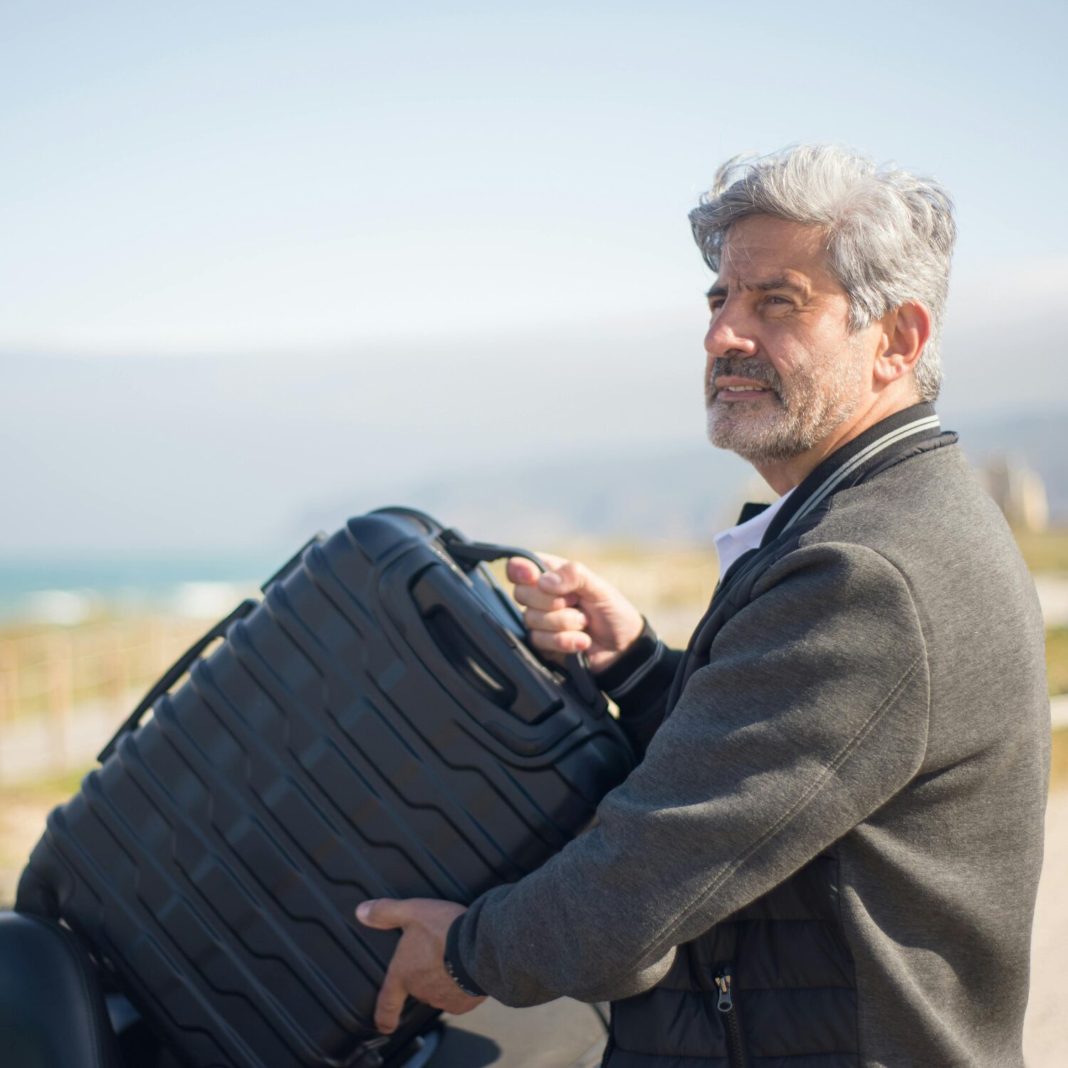 An Elderly Caucasian man wearing a gray jacket carrying a black travel luggage outdoors.