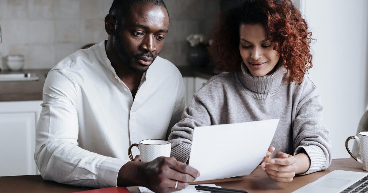 315 A Black man wearing a white long sleeved blouse and a Black woman wearing a grey turtleneck sweater, sitting beside each other while reading a document.