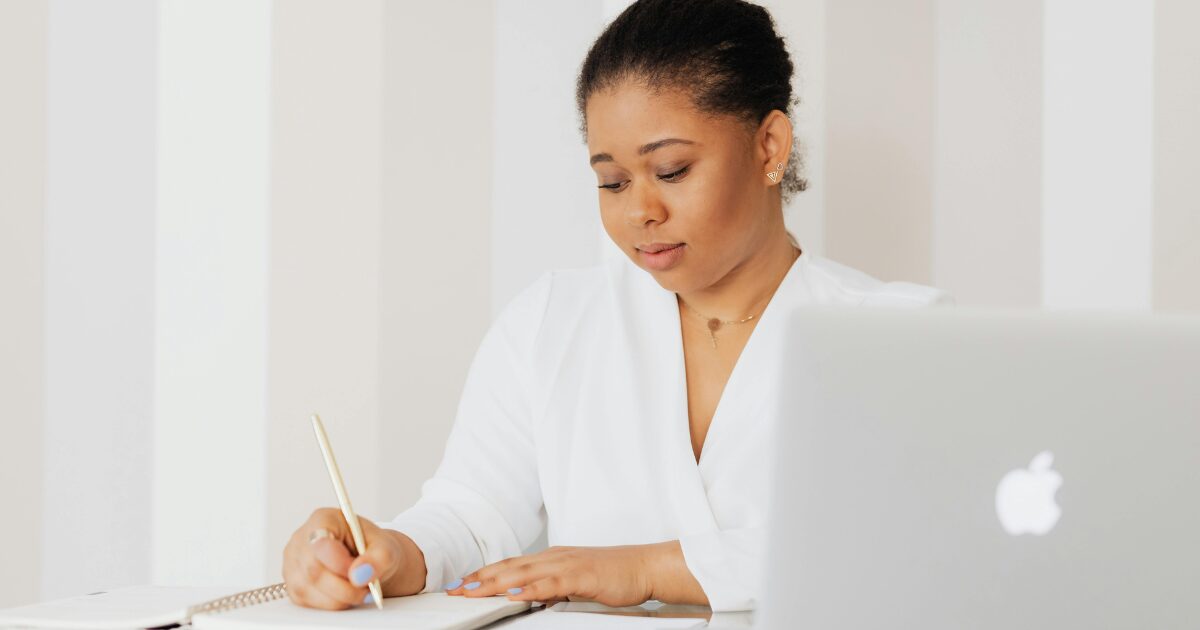 A Black woman wearing a white blouse writing on a document while sitting in front of a laptop