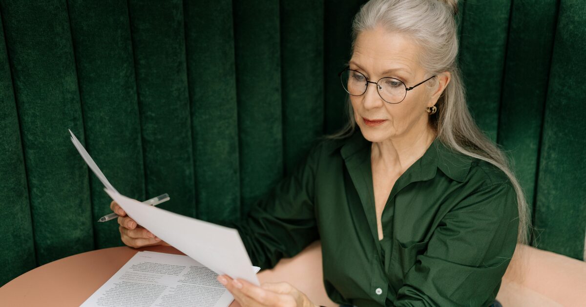 In-Blog Thumbnails An Elderly Caucasian woman wearing a dark green blouse holding documents and a pen on her right hand.