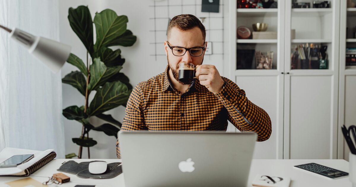 A Caucasian man wearing an orange checkered shirt sipping on a cup while looking at a laptop